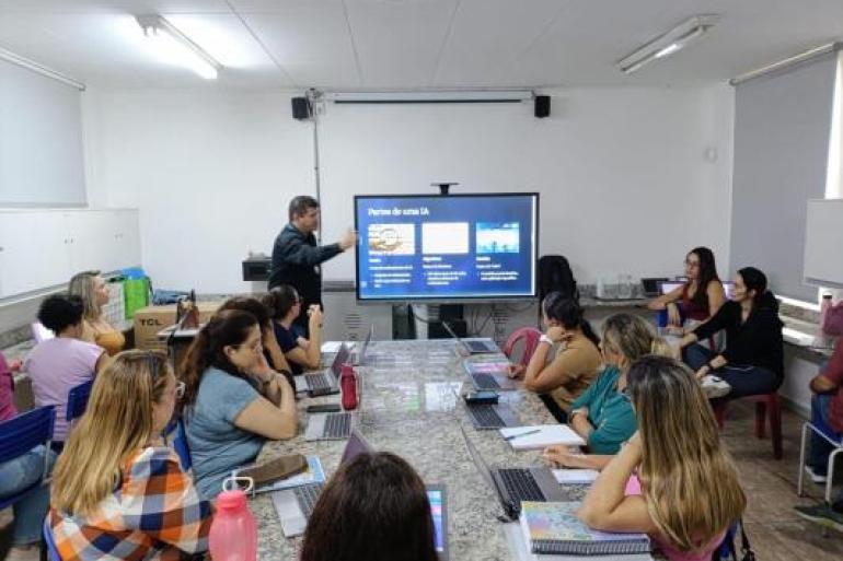 Formação reuniu professores dos Anos Finais do Ensino Fundamental #paratodosverem: Educador apresenta conteúdo sobre IA em sala de aula, usando uma tela interativa. Os demais professores, sentados ao redor de uma mesa com laptops e cadernos, acompanham atentos e participam da discussão em um ambiente colaborativo de aprendizagem.