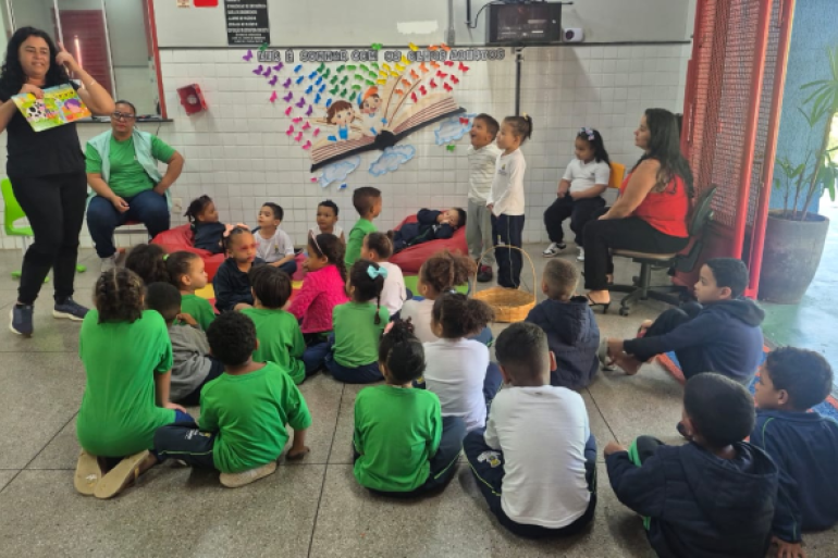#PARATODOSVEREM: Professora conduz leitura para grupo de crianças sentadas em roda, em espaço colorido e decorado, acompanhadas por outras educadoras. 