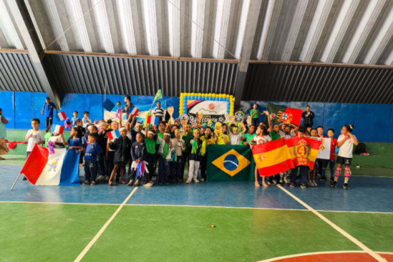 #paratodosverem: Um grande grupo de crianças posa reunido em uma quadra poliesportiva de piso verde e azul, sob teto metálico. Elas seguram bandeiras de diferentes países, como Brasil, França, Espanha e Portugal, além de erguerem troféus. Ao fundo, há uma parede azul e um painel emoldurado por balões amarelos e azuis, caracterizando uma gincana escolar.