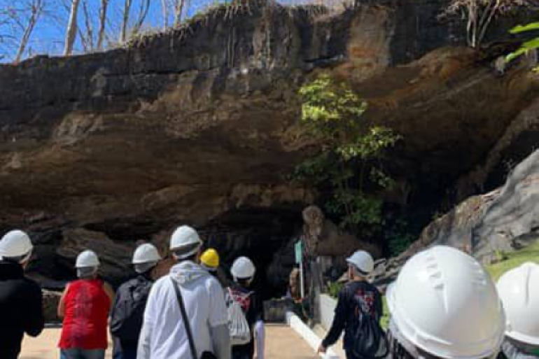 Visita técnica leva estudantes a conhecerem formações rochosas e processos naturais. #paratodosverem: Estudantes, vistos de costas, caminham em direção a uma formação rochosa ao ar livre. Eles usam capacetes de segurança e estão acompanhados em uma área que parece um sítio geológico.