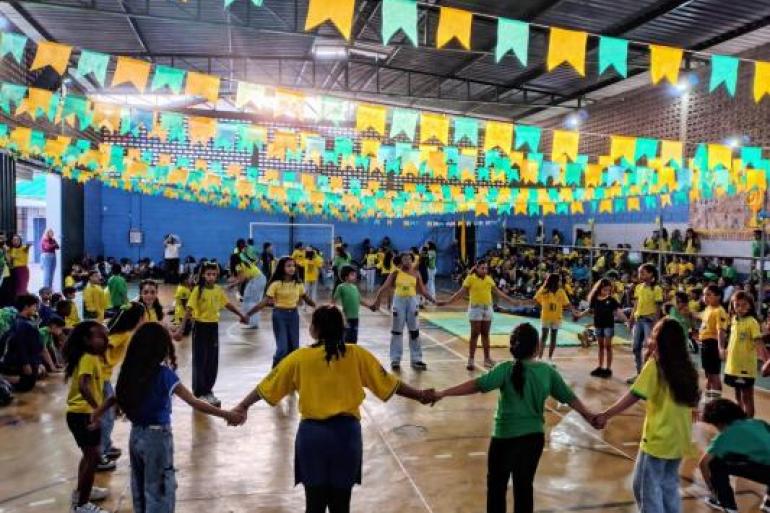 Estudantes participam de apresentação durante evento escolar #paratodosverem: Em quadra coberta, estudantes participam de uma atividade, vestidos com roupas nas cores verde e amarelo. No centro, eles formam uma roda de mãos dadas. O ambiente está decorado com bandeirinhas juninas também das cores verde e amarelo no teto, enquanto outros alunos acompanham a programação ao redor da quadra.