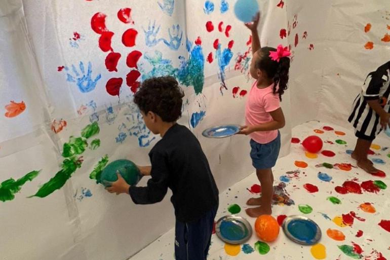 #paratodosverem: A imagem mostra três crianças brincando em um espaço coberto por papel branco no chão e paredes. Elas seguram balões mergulhados em tinta colorida e os usam para pintar o papel, criando marcas de mãos e pintura. O chão está cheio de pratos metálicos com tinta de várias cores, como rosa, azul e amarelo.