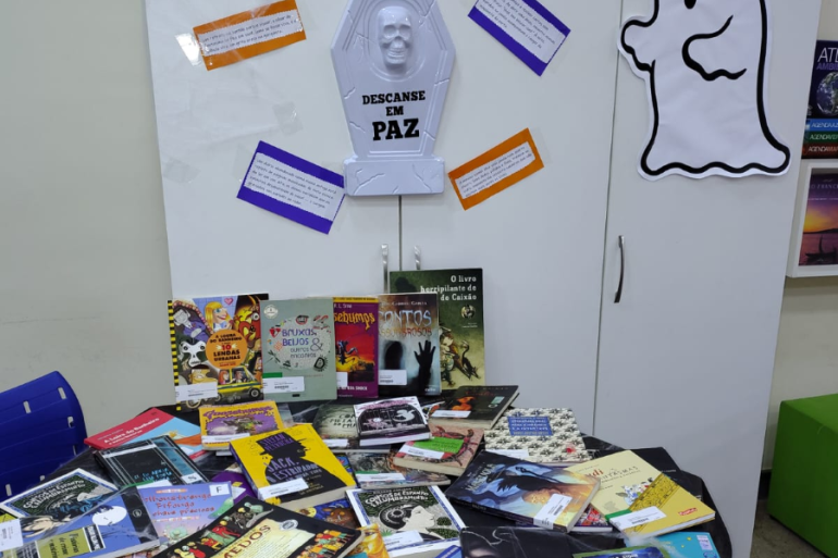 Escola faz atividade temática de suspense e terror #paratodosverem: A imagem mostra uma exposição temática de livros intitulada “Beco do Arrepio”, montada em um ambiente escolar. A decoração inclui elementos que remetem ao Halloween, como uma lápide com os dizeres “Descanse em Paz”, teias de aranha, aranhas de brinquedo e a figura de um fantasma na parede. Sobre a mesa, há uma grande variedade de livros de mistério, terror e suspense.