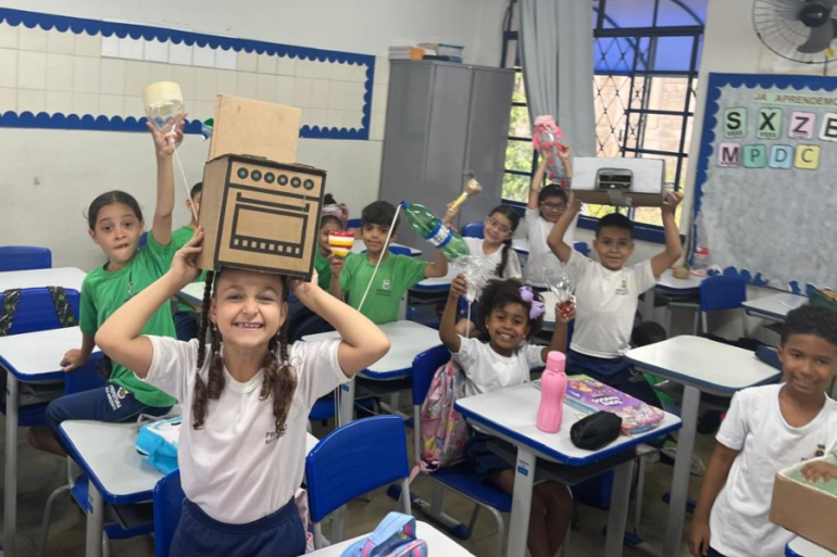 #paratodosverem: Em uma sala de aula iluminada, um grupo de crianças está animado em suas carteiras, segurando objetos reciclados acima da cabeça. Uma menina no centro ri, usando uma caixa de papelão desenhada como fogão.