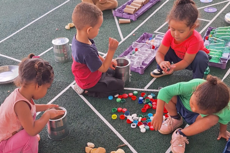 #paratodosverem: Quatro crianças estão sentadas no chão de um pátio, que tem marcações esportivas, brincando com materiais reciclados. Elas interagem com latas de metal, garrafas plásticas, papelão, pedras e tampinhas coloridas espalhadas pelo chão, em uma atividade lúdica que incentiva a criatividade e o uso de materiais não estruturados