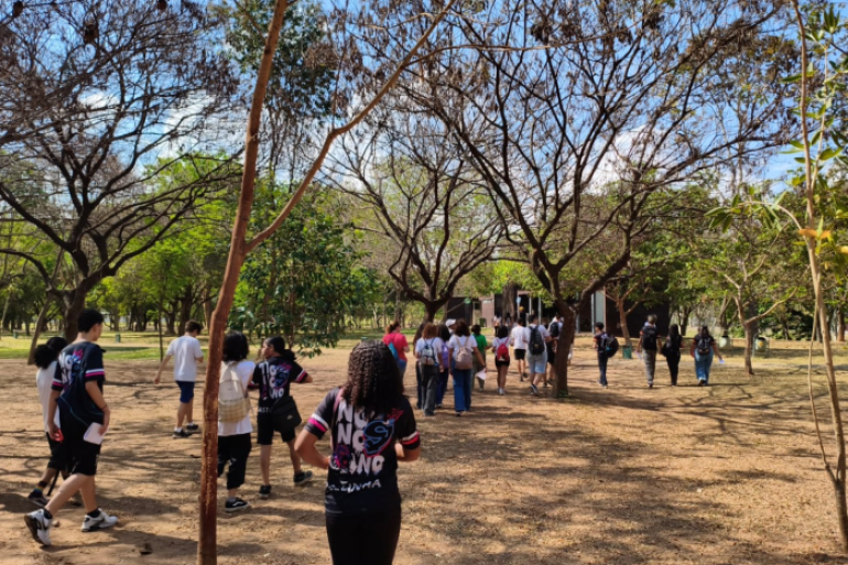 #paratodosverem: Um grupo de jovens, a maioria de costas, caminha em uma trilha de terra sob a copa de árvores secas e frondosas, em um parque. A cena, vista por trás de um tronco, mostra os participantes de mochilas, em um momento de lazer e aprendizado ao ar livre.