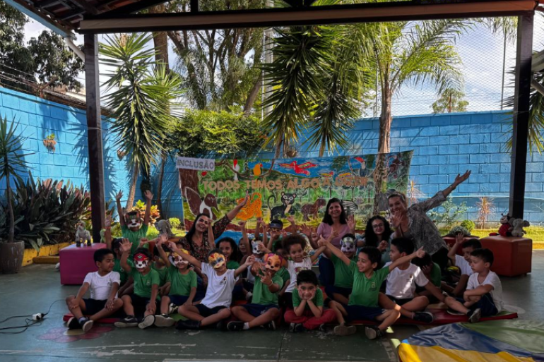 Peça teatral tem personagem que se comunica por Comunicação Alternativa Aumentativa (CAA) #paratodosverem: Crianças e educadoras posam sorrindo em um pátio aberto, muitas delas usando pintura facial de animais. Atrás, um painel colorido com desenhos da fauna e a frase “Todos temos algo a oferecer”.