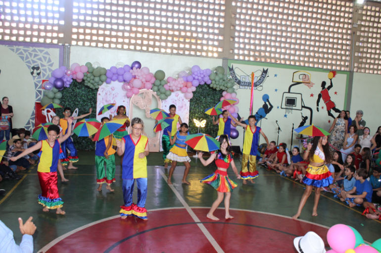 Cores, dança e alegria celebram a infância em cena cultural #paratodosverem: Na foto, crianças se apresentam em um ginásio escolar decorado com balões coloridos. Elas usam fantasias vibrantes em azul, amarelo, vermelho e verde, e seguram sombrinhas multicoloridas, típicas de danças populares brasileiras. Com sorrisos e movimentos coreografados, enchem o espaço de cor e alegria, enquanto famílias assistem animadas ao espetáculo.