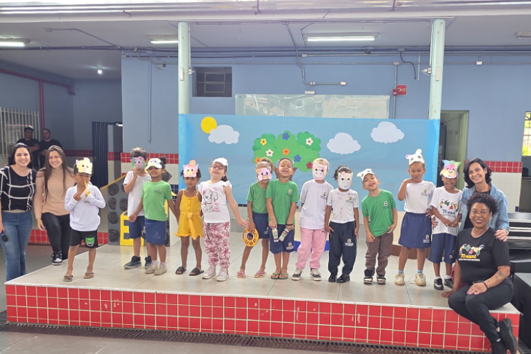 Aprendizado e diversão no palco: arte e teatro em cena Legenda: Aprendizado e diversão no palco: arte e teatro em cena #paratodosverem: A imagem mostra um grupo de crianças em um palco decorado com um painel azul, nuvens, sol e árvores de papel. As crianças usam máscaras de porquinhos e roupas coloridas. À frente do palco, algumas crianças estão sentadas no chão sobre um tapete colorido com números. Quatro professoras sorriem junto ao grupo.