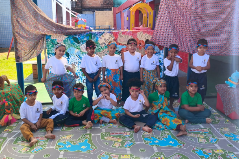 Celebrando a cultura afro-brasileira e indígena com aprendizado, cores e ancestralidade #paratodosverem: Na imagem, um grupo de crianças posa sorridente em um espaço decorado com tecidos coloridos, representando um cenário cultural. Elas usam faixas na cabeça e roupas estampadas, sugerindo uma atividade escolar temática, ligada à cultura afro-brasileira e indígena
