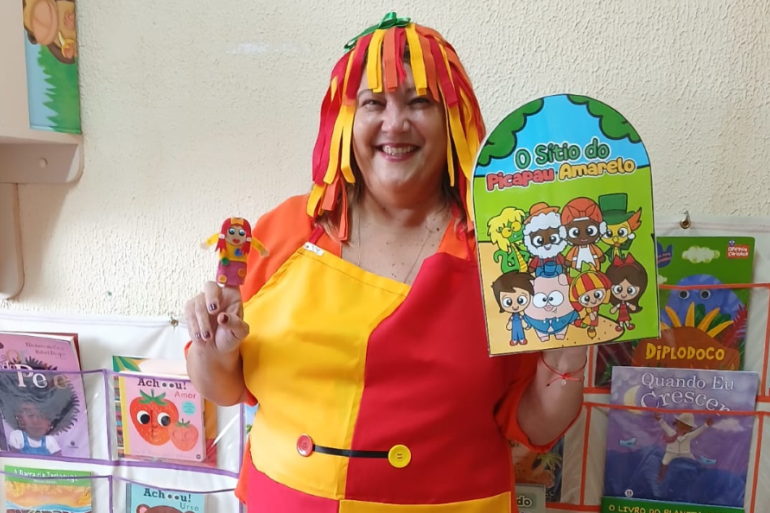 Com leituras divertidas, professora inspira crianças a mergulharem na fantasia #paratodosverem: Na foto, uma mulher sorri, vestida como a personagem Emília, do Sítio do Picapau Amarelo. Ela usa uma fantasia colorida em vermelho e amarelo, com uma peruca de tiras coloridas. Em uma mão segura um livrinho do Sítio e, na outra, uma pequena boneca. Ao fundo, há prateleiras cheias de livros infantis.