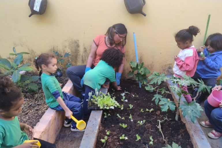#paratodosverem: A imagem mostra um grupo de crianças pequenas participando de uma atividade de horta escolar. Elas estão acompanhadas por uma adulta, que orienta o plantio em um canteiro de terra. Algumas crianças seguram ferramentas de jardinagem e regadores, enquanto outras observam e ajudam a plantar mudas.