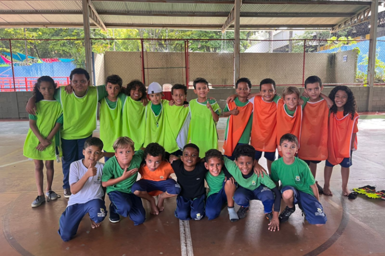 #paratodosverem: Em uma quadra coberta, um grupo grande de crianças, vestindo coletes verde-limão e laranja, está posando para uma foto em duas fileiras. Eles estão abraçados, com sorrisos e gestos de "joinha". 