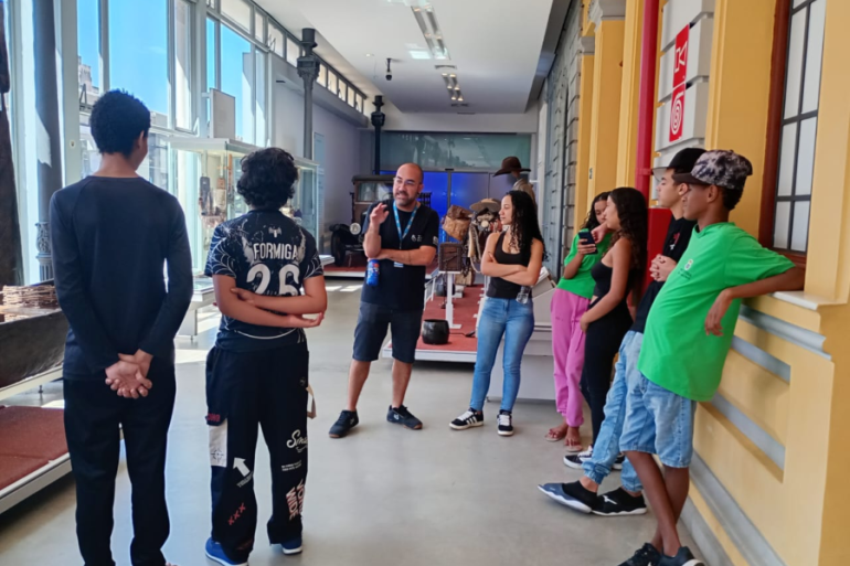 #paratodosverem: Em um corredor de museu, iluminado por grandes janelas laterais, um grupo de jovens está reunido em torno de um professor ou monitor. O monitor, de camiseta preta e crachá, gesticula enquanto conversa com o grupo. A cena, com pisos claros e paredes amarelas, transmite um momento de aprendizado e visita cultural.