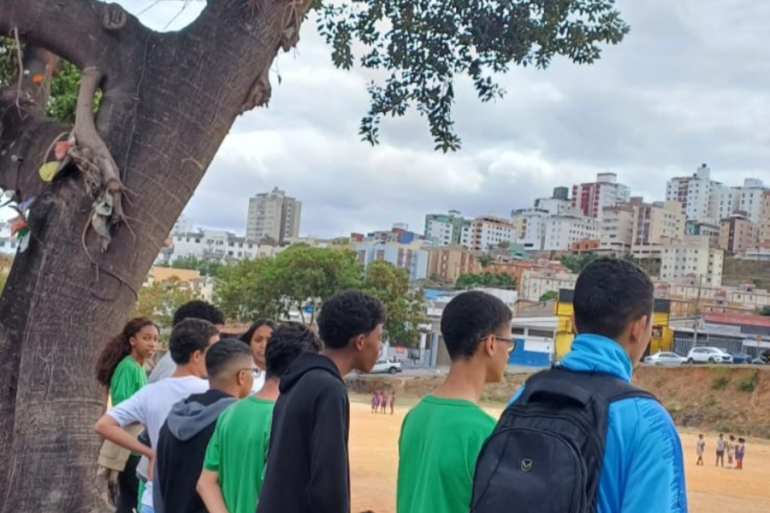 #paratodosverem: Um grupo de jovens, em sua maioria meninos, está de pé em uma área de terra, olhando para um campo de futebol abaixo e para uma paisagem urbana com muitas casas e prédios em um morro ao longe. A cena é em um dia nublado. Em primeiro plano, um tronco de árvore retorcido enquadra a vista da comunidade