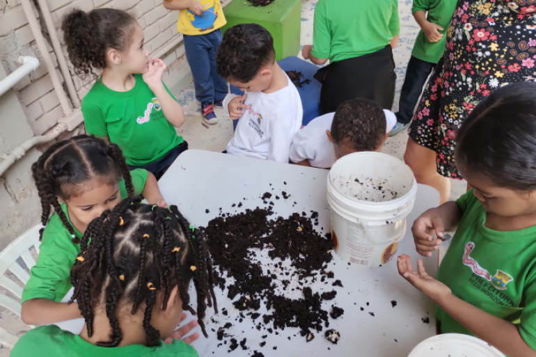 #paratodosverem: Crianças pequenas participam de uma atividade prática em grupo, explorando o conteúdo de baldes com terra e minhocas sobre uma mesa. Elas usam camisetas verdes com o logotipo da Prefeitura de Belo Horizonte.
