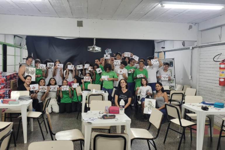 #paratodosverem: Um grupo de estudantes posa em uma sala de aula segurando trabalhos feitos com folhas e elementos naturais. Eles sorriem e mostram suas produções. Algumas professoras estão sentadas na frente, também segurando os trabalhos. Mesas e cadeiras estão espalhadas pelo espaço.