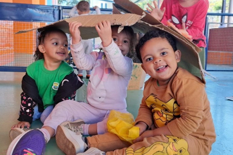 Estudantes exploram papelão a partir de livro sobre reciclagem #paratodosverem: Três crianças pequenas estão sentadas no chão de um pátio, rindo e brincando sob um pedaço de papelão que usam como cabana. A menina da esquerda veste uma camiseta verde, a do meio veste rosa claro, e o menino da direita veste uma blusa laranja com desenho do Rei Leão.