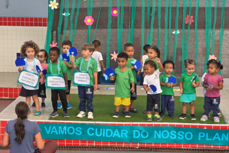 #paratodosverem: Um grupo de 15 crianças, a maioria em idade pré-escolar e vestindo uniformes verdes, está em um palco. Três crianças seguram cartazes com frases sobre a natureza, como "VAMOS CUIDAR DA NATUREZA". Ao fundo, o cenário é decorado com flores de papel. Em primeiro plano, uma professora está sentada no chão, de costas, em frente a uma faixa azul com texto.