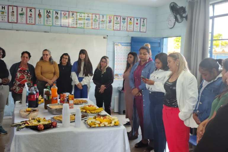 Encontro visou a discutir sobre a educação inclusiva #paratodosverem: Em uma sala de aula decorada com o alfabeto, um grupo de professoras e funcionárias está reunido ao redor de uma mesa com salgados, frutas e refrigerantes. A maioria das mulheres está de pé, conversando e atenta.
