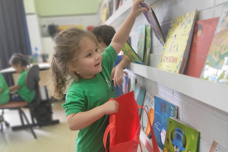 #paratodosverem: A imagem mostra uma criança pequena sorridente, usando camiseta verde, escolhendo um livro em uma estante cheia de títulos infantis. Ela segura uma sacola vermelha aberta, como se fosse guardar o livro dentro dela. Ao fundo, outras crianças também estão ocupadas na sala de aula.