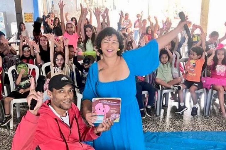 #paratodosverem: Em um espaço interno de uma escola, um homem em cadeira de rodas segurando um livro e uma mulher vestindo um vestido azul com um microfone sorriem enquanto estão à frente de um grupo de crianças sentadas em cadeiras de plástico. Todos estão levantando as mãos. Algumas crianças usam fantasias coloridas.