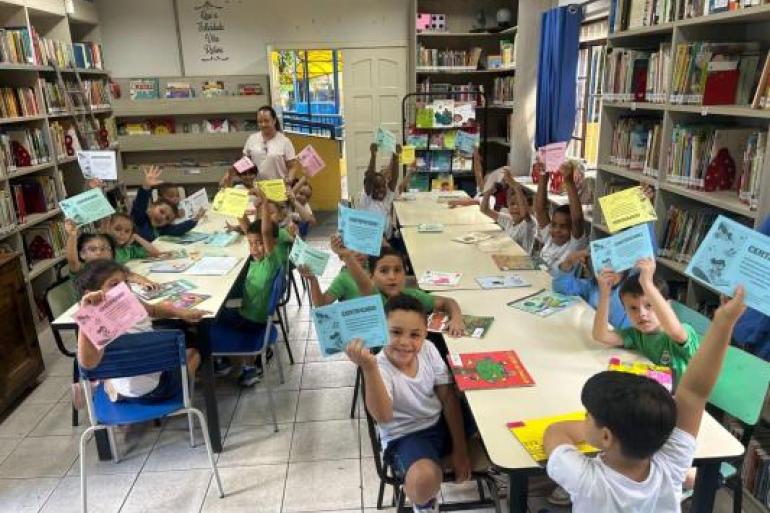 #paratodosverem: Crianças sentadas, em biblioteca escolar, mostram certificados de diferentes cores enquanto sorriem ao redor de mesas com livros e atividades. Ao fundo, há estantes com livros e uma educadora acompanha o grupo.
