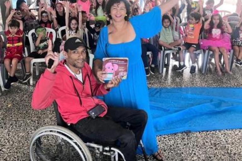 #paratodosverem: Em um espaço interno de uma escola, um homem em cadeira de rodas segurando um livro e uma mulher vestindo um vestido azul com um microfone sorriem enquanto estão à frente de um grupo de crianças sentadas em cadeiras de plástico. Todos estão levantando as mãos. Algumas crianças usam fantasias coloridas.