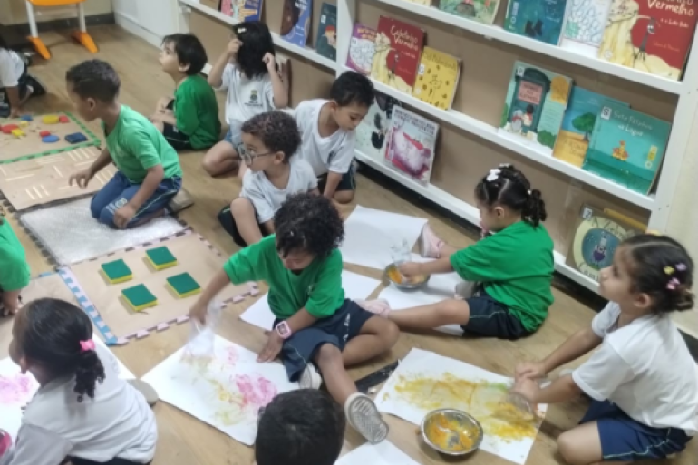Escola promoveu atividades para fortalecer o vínculo com famílias #paratodosverem: Crianças pequenas sentadas no chão de madeira de uma sala de aula participam de uma atividade artística. Elas usam potes com tinta amarela e rosa para pintar em grandes folhas de papel espalhadas pelo chão. Algumas usam uniforme verde e outras, branco. Ao fundo, uma estante branca exibe diversos livros infantis virados para a frente, e há tapetes sensoriais dispostos à esquerda.