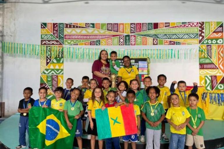 Mini Copa promove integração e aprendizado entre estudantes #paratodosverem: Várias crianças posam para uma foto vestidas com as cores verde e amarelo e com medalhas no pescoço. Na frente, duas crianças seguram as bandeiras do Brasil e de Senegal. Ao fundo, duas professoras acompanham o grupo, com um troféu erguido por um dos estudantes.