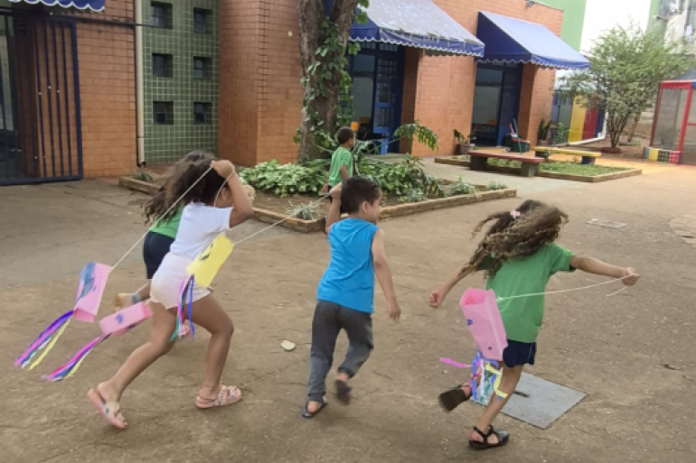 Os alunos são incentivados a descobrir os brinquedos #paratodosverem: Cinco crianças correm em um pátio escolar de terra, brincando com rabiolas coloridas presas a cordões que seguram pelas mãos. Três delas correm para a direita, com fitas rosa, amarela e coloridas voando atrás. Ao fundo, há um edifício com tijolos à vista e toldos azuis sobre portas e janelas. Canteiros com plantas verdes e um banco de madeira também estão visíveis.