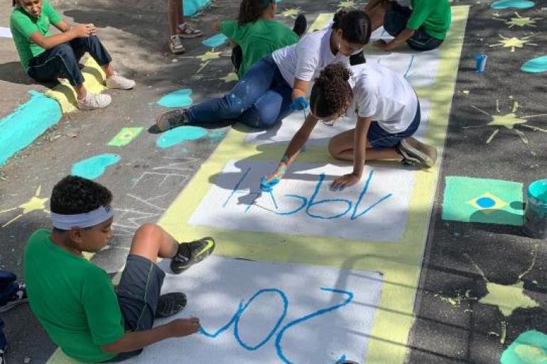 #paratodosverem: Estudantes participam da pintura de uma rua decorada para a Copa do Mundo. Sentados no chão, eles utilizam tinta azul para escrever em espaços demarcados”. Ao redor, aparecem desenhos de estrelas, bandeiras do Brasil e círculos em verde, amarelo e azul.