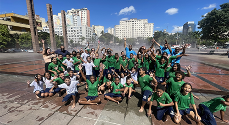 na imagem, um grupo grande de crianças e pessoas adultas está reunido em um espaço público ao ar livre, com roupas molhadas e expressões de alegria.