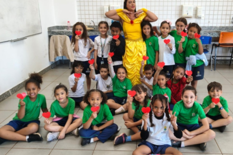 A importância de falar sobre os sentimentos foi um dos pilares do projeto. #paratodosverem: Diversas crianças de uniforme escolar posam sentadas e em pé em uma sala de aula iluminada. No centro, uma mulher vestida como a princesa Bela, de vestido amarelo, faz uma pose divertida. A maioria das crianças segura pequenos corações de papel vermelho em direção à câmera, sorrindo. Ao fundo, há um quadro branco, uma bancada de granito com pia e cadeiras organizadas na sala.