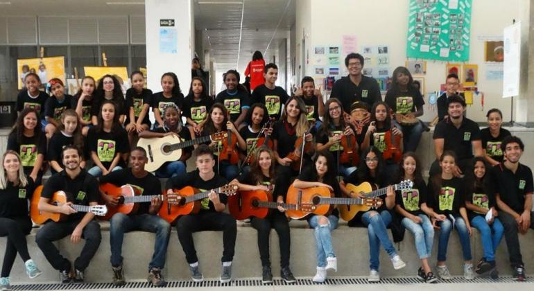 Programa Música na Escola Cerca de 30 pessoas, entre alunos e professores, estão sentados em uma arquibancada, usando camisetas pretas, ealgumas delas seguram violões