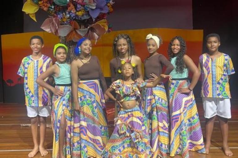 #paratodosverem: Grupo de oito crianças usando figurinos coloridos em diferentes tons de cores. Ao fundo, há um cenário com uma grande árvore de flores multicoloridas e painéis em degradê de laranja e vermelho.