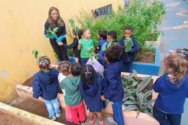#paratodosverem: Uma professora de óculos, roupa e calça pretas e luvas azuis segura folhas de hortaliça enquanto orienta um grupo de 12 crianças pequenas ao seu redor. Diversos alunos também seguram folhas e observam a atividade. Ao fundo e nas laterais, há canteiros com plantas verdes e o pátio da escola. 