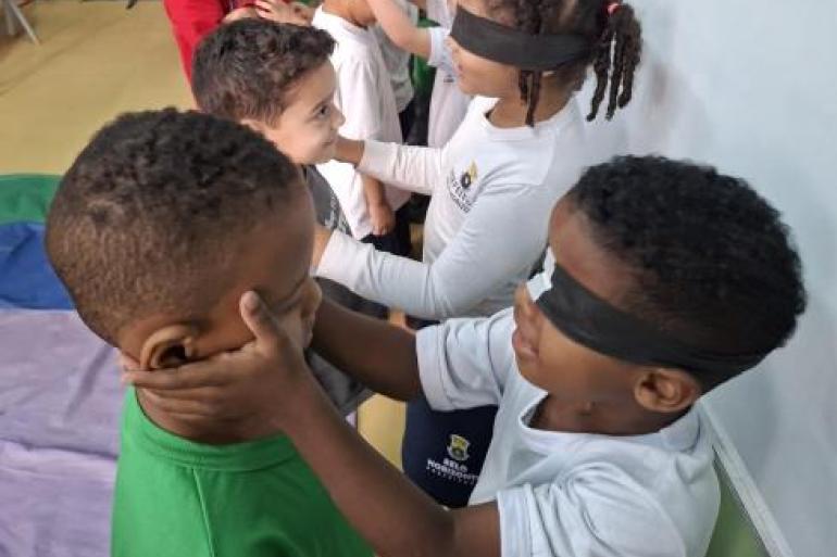 Crianças participam de brincadeira com as mãos #paratodosverem: Em uma sala de aula, crianças participam de uma atividade sensorial em duplas. Em primeiro plano, uma criança vendada, vestindo camiseta branca, toca delicadamente com as mãos o rosto de outra criança com camiseta verde, utilizando as mãos para reconhecer seus traços. Ao fundo, outras duplas realizam a mesma dinâmica, também com algumas crianças vendadas.