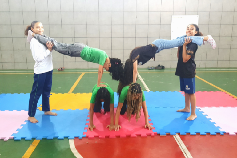 #paratodosverem: Em uma quadra esportiva com piso nas cores verde e vermelho, seis jovens realizam uma formação acrobática. No centro, duas estudantes estão de joelhos e mãos apoiadas sobre tatames coloridos de encaixe (nas cores azul, rosa e amarelo). Sobre as costas delas, outras duas fazem paradas de mão, tendo as pernas seguradas horizontalmente por duas jovens em pé nas laterais. Todas vestem uniformes.
