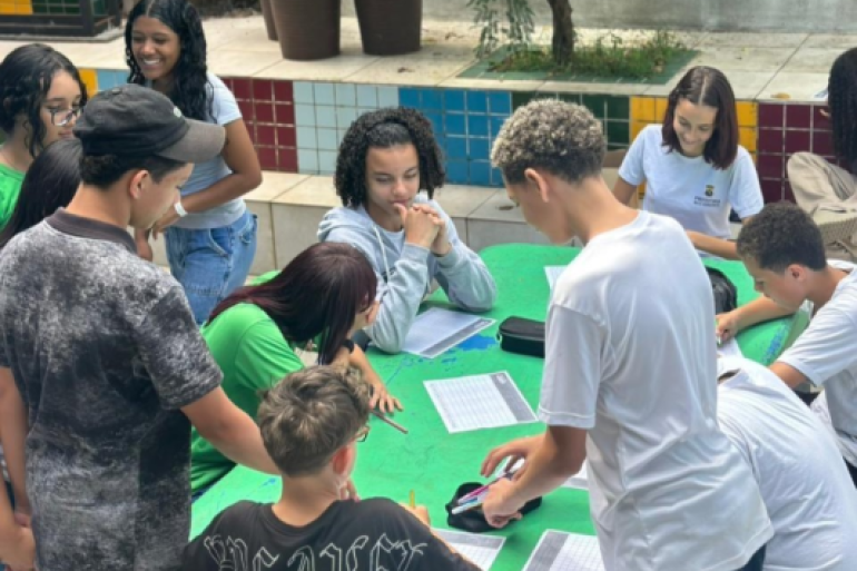 #paratodosverem: Em um pátio externo, cerca de 12 jovens (estudantes) estão reunidos em torno de uma mesa de cimento pintada de verde. Eles estão sentados e em pé, escrevendo em folhas de papel com grades. Alguns vestem camisetas de uniformes verdes e brancas. Há estojos e canetas sobre a mesa. Ao fundo, veem-se bancos com azulejos coloridos (vermelho, azul, verde, amarelo) e vasos de plantas.