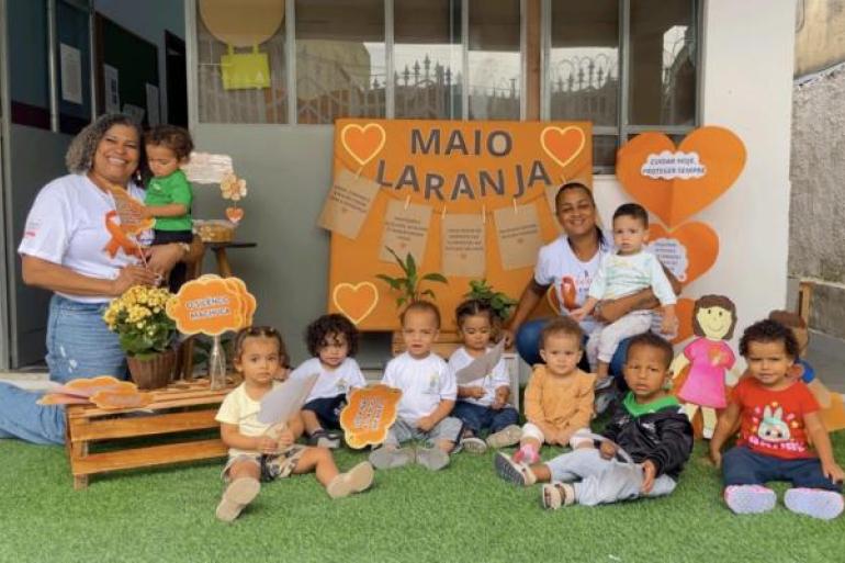 #paratodosverem: Crianças e educadoras participam de atividade do Maio Laranja em espaço escolar decorado com cartazes e corações na cor laranja. Ao fundo há mensagens de conscientização sobre proteção da infância, enquanto os participantes posam sentados sobre gramado sintético. 