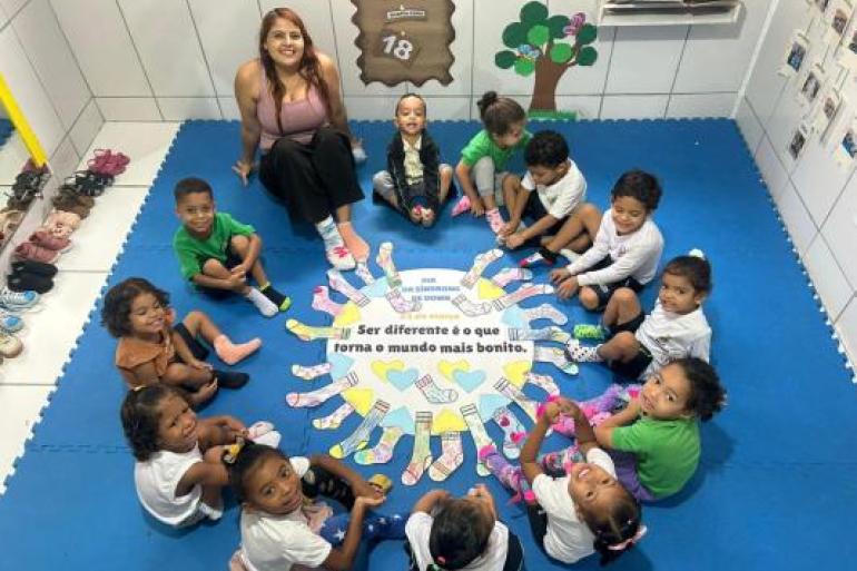 #paratodosverem: Crianças e uma professora sentadas em círculo sobre um tapete azul participam de atividade em sala. No centro, há um cartaz sobre o Dia da Síndrome de Down, com desenhos de meias coloridas e de corações nas cores azul e amarela, além da frase: “Ser diferente é o que torna o mundo mais bonito”.