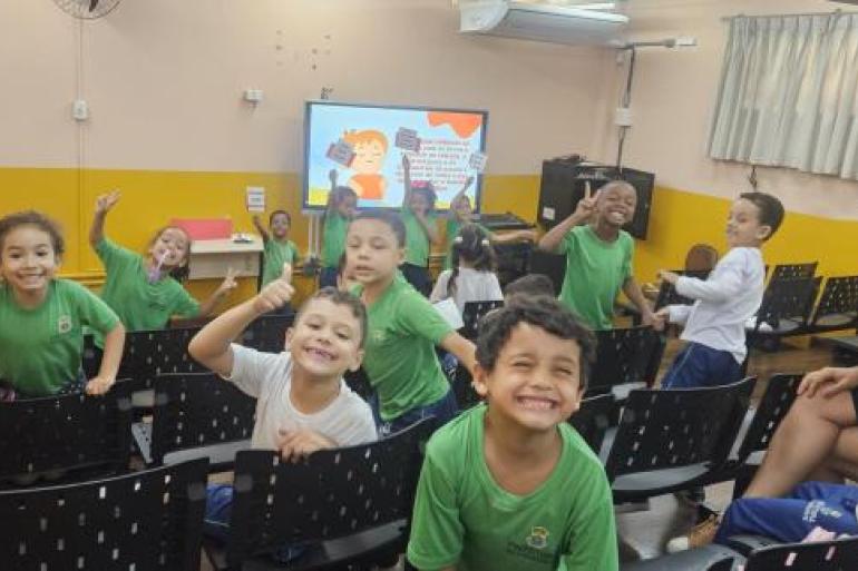 #paratodosverem: Em uma sala com cadeiras pretas organizadas em fileiras, várias crianças usam uniformes escolares verdes e brancos e sorriem para a câmera. Outras crianças também uniformizadas seguram pequenas plaquinhas com frases de cuidado aos livros. À direita, uma professora de vestido preto e óculos sorri sentada em uma das cadeiras. Ao fundo, há uma televisor com uma imagem projetada
