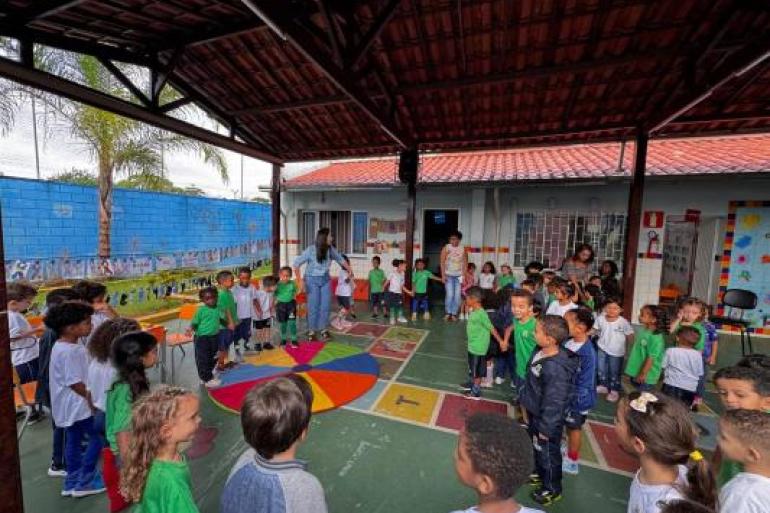 #paratodosverem: Crianças organizadas em roda participam de uma atividade coletiva em pátio escolar coberto, orientada por professores. No chão, há um tapete circular colorido e marcações de jogos. Ao fundo, aparecem murais com trabalhos feitos pelos alunos.