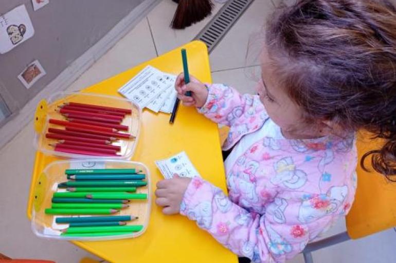 Criança avalia refeição do dia #paratodosverem: Em uma mesa amarela, uma criança segura um lápis enquanto avalia a refeição oferecida pela creche. Sobre a mesa, há cartões ilustrados e recipientes com lápis das cores vermelha e verde.