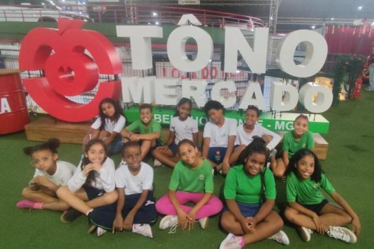 #paratodosverem: Grupo de crianças posa sentado à frente do letreiro “TÔ NO MERCADO”, em um espaço com estrutura metálica e piso verde. Os alunos usam uniforme escolar com blusas nas cores branca e verde e sorriem para a foto durante a visita ao Mercado.