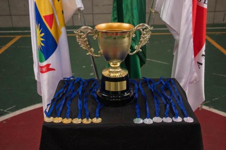 #paratodosverem: Troféu dourado está sobre uma mesa coberta por pano preto em uma quadra esportiva. Ao lado, há fileiras de medalhas com fitas azuis, nas cores dourada e prata. Ao fundo, aparecem as bandeiras do Brasil e de Minas Gerais.