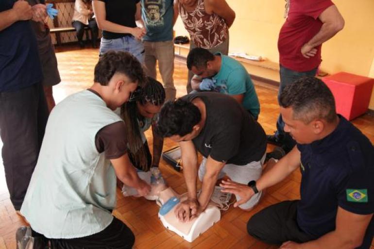 #paratodosverem: Grupo de adultos participa de um treinamento de primeiros socorros em sala. No centro, um homem faz compressões no peito de um boneco de treino, enquanto outras pessoas observam e ajudam.