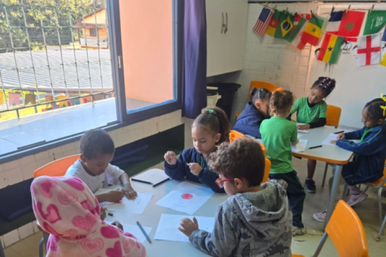 Estudantes desenharam bandeiras dos países e bolas de futebol. #paratodosverem: Crianças pequenas sentadas em mesas brancas com cadeiras laranja desenham a bandeira do Japão em folhas de papel numa sala de aula. À esquerda, uma janela com grade de ferro preta e cortina roxa. Na parede do fundo, um varal com bandeiras de vários países, incluindo Brasil, EUA, Camarões e Senegal.
