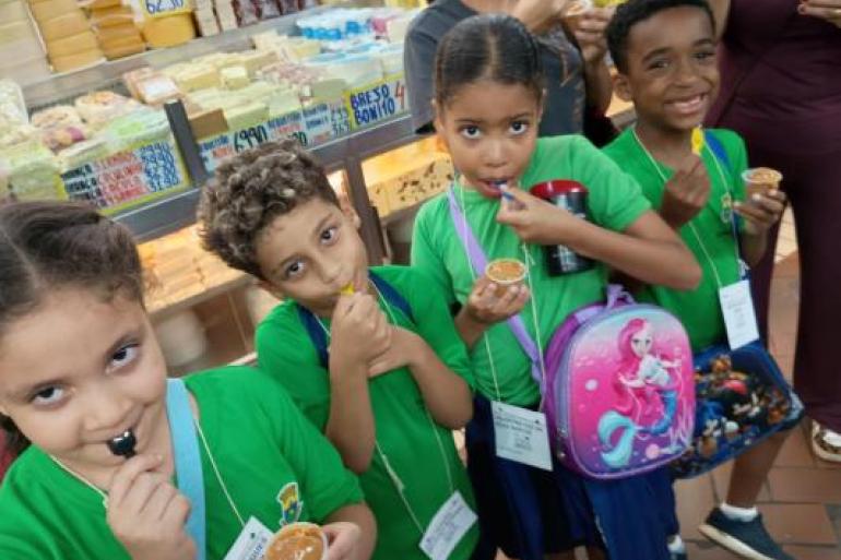 #paratodosverem: Quatro crianças, dois meninos e duas crianças, usando uniformes escolares da Prefeitura de Belo Horizonte e crachás, provam doce no Mercado Central. Cada uma segura um pequeno copo com doce de leite e colher, enquanto ao fundo aparecem balcões refrigerados com queijos, etiquetas de preços e produtos expostos